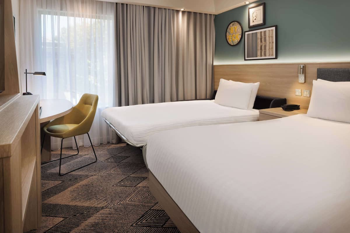 A hotel room with two beds, a desk, a chair, and a large window with curtains.
