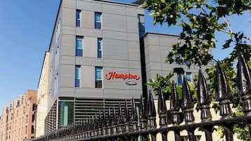 Un bâtiment moderne, orné d’une enseigne « Hampton by Hilton », avec une façade géométrique et une clôture métallique en premier plan.