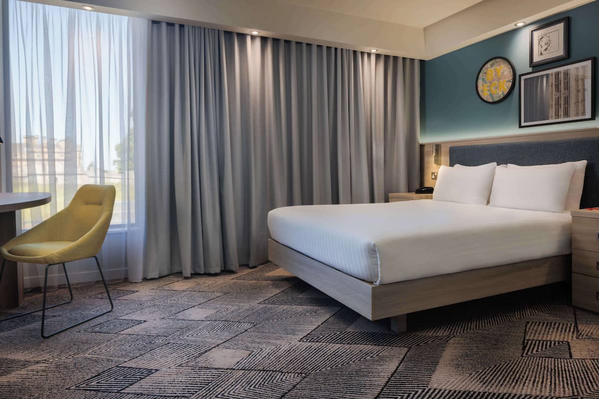 A hotel room with a bed, a chair, a desk, and a large window with curtains.