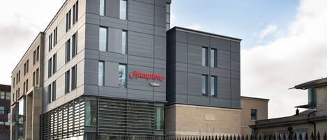 A modern building with a 'Hampton' sign, featuring a flat facade, large windows, and a metal fence in front.
