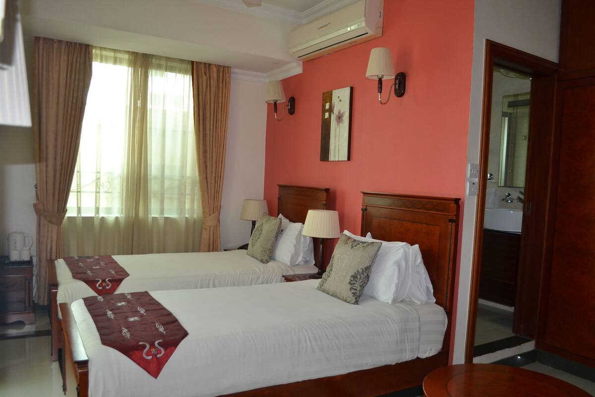 Quarto de hotel com duas camas, cabeceira de madeira, mesa de cabeceira, luminária, espelho e janela com cortinas.