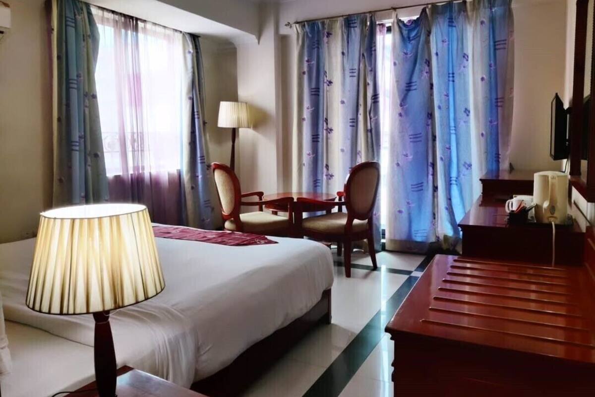 Quarto de hotel com cama, escrivaninha, cadeira, abajur e cortinas.