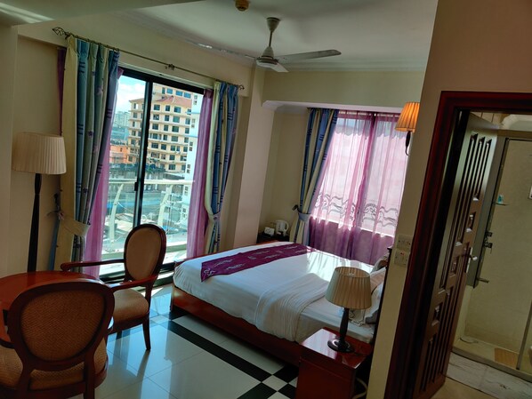 Habitación de hotel con cama, una silla, una mesa, lámpara y vistas a edificios a través del ventanal.