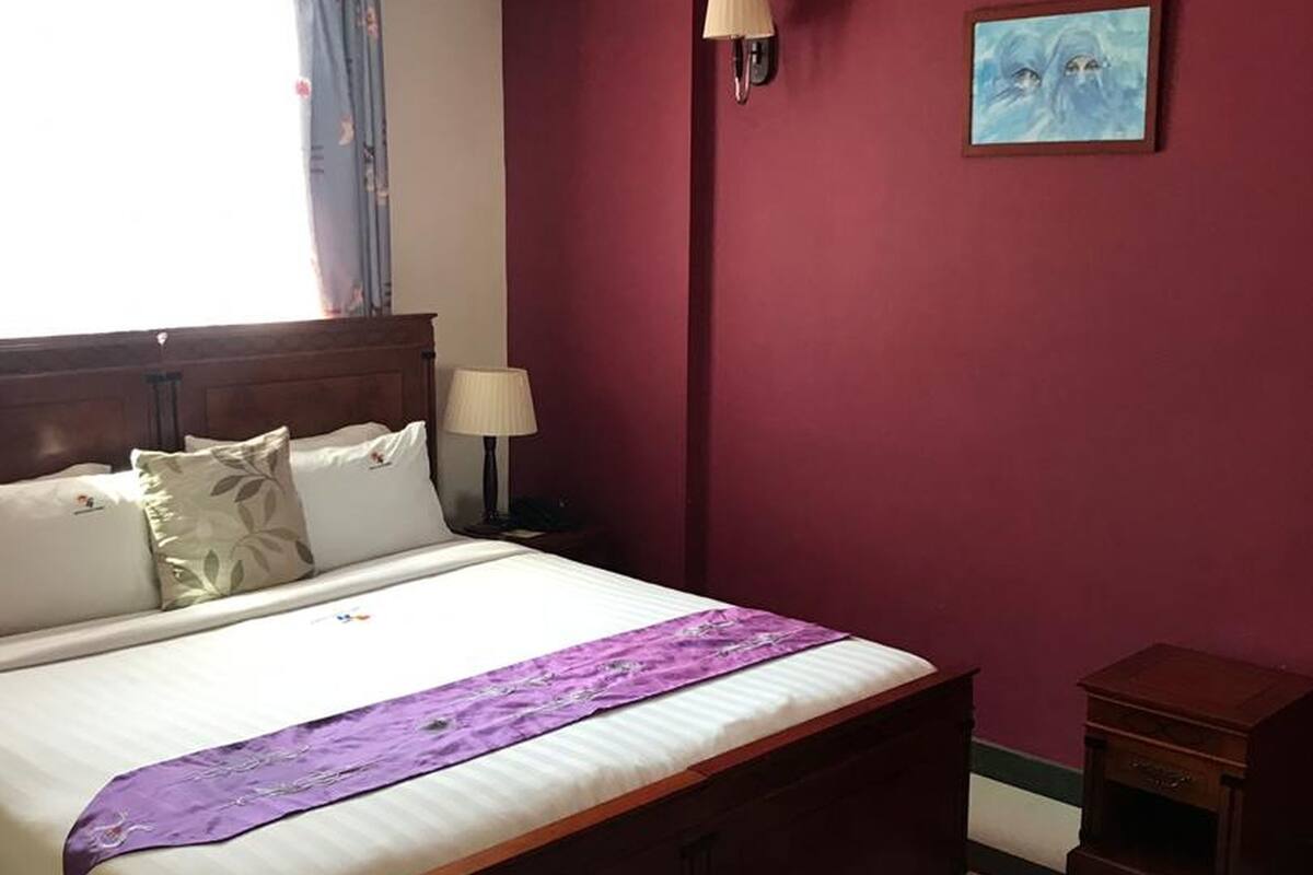 Quarto com cama, mesa de cabeceira, luminária, pintura e janela com cortinas.