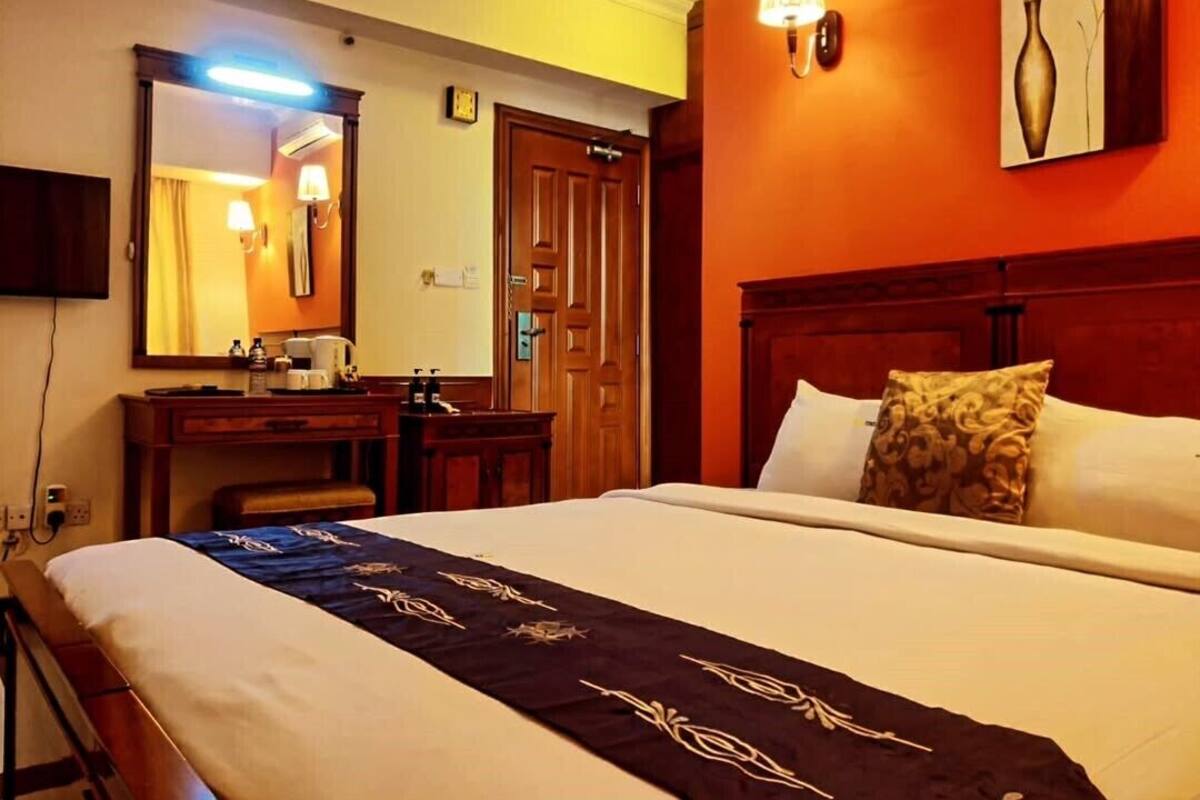 Quarto de hotel com uma cama grande, uma escrivaninha com cadeira, um espelho, uma luminária na parede e uma porta.