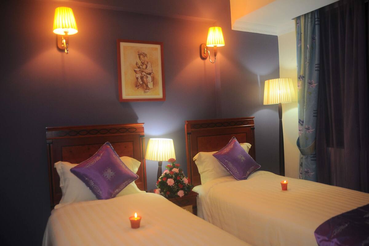 Quarto de hotel com duas camas, cada uma com uma almofada roxa, uma mesa de cabeceira com abajur, um quadro na parede e um arranjo floral.