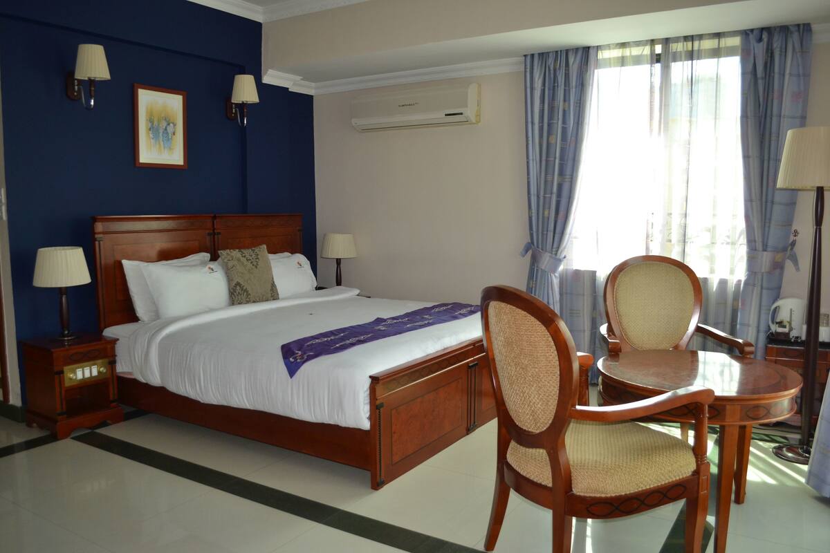 Quarto de hotel com uma cama grande, duas mesas de cabeceira com luminárias, uma cadeira de madeira, uma pequena mesa redonda e uma janela com cortinas.