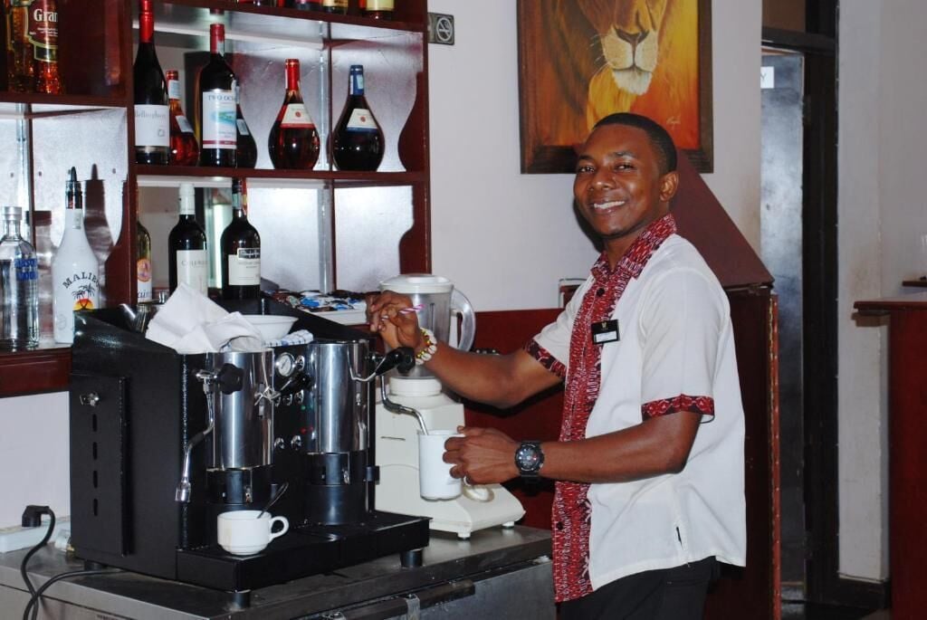 Un barista preparando una bebida en una máquina de café en un bar, con estantes llenos de botellas al fondo.