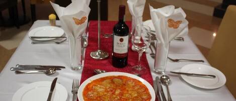 A dining table set with a red tablecloth, white plates, silverware, and a bottle of wine.