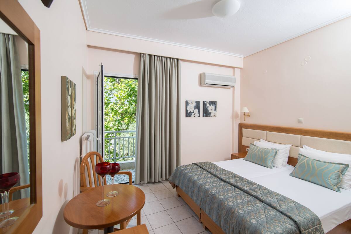 Habitación de hotel con cama, una mesa de comedor con sillas, ventana con cortinas y obras de arte en las paredes.