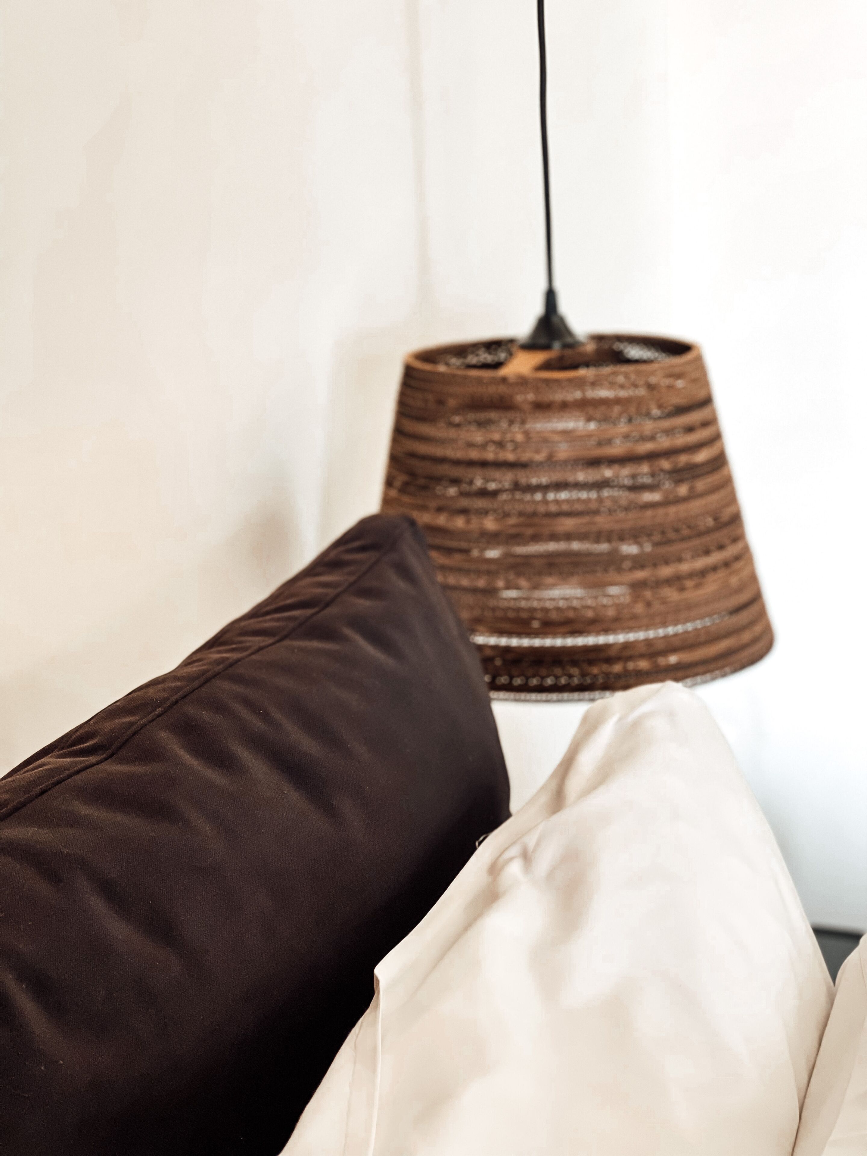 A room with a hanging woven lamp and two pillows, one dark and one light, on a white wall.