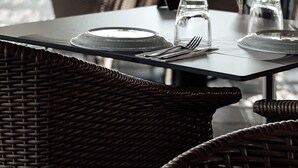 A dining area with wicker chairs and tables, clear glassware, and a view of trees and a building in the background. - Dion Palace Resort & Spa by Anayia All Inclusive Resorts (Dio-Olympos)