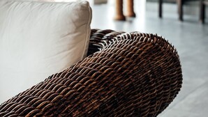 A wicker chair with a white pillow in a modern indoor space with large windows and a bar area in the background. - Dion Palace Resort & Spa by Anayia All Inclusive Resorts (Dio-Olympos)