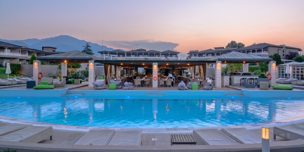 A pool area with lounge chairs and a covered seating area, surrounded by buildings with a view of mountains.