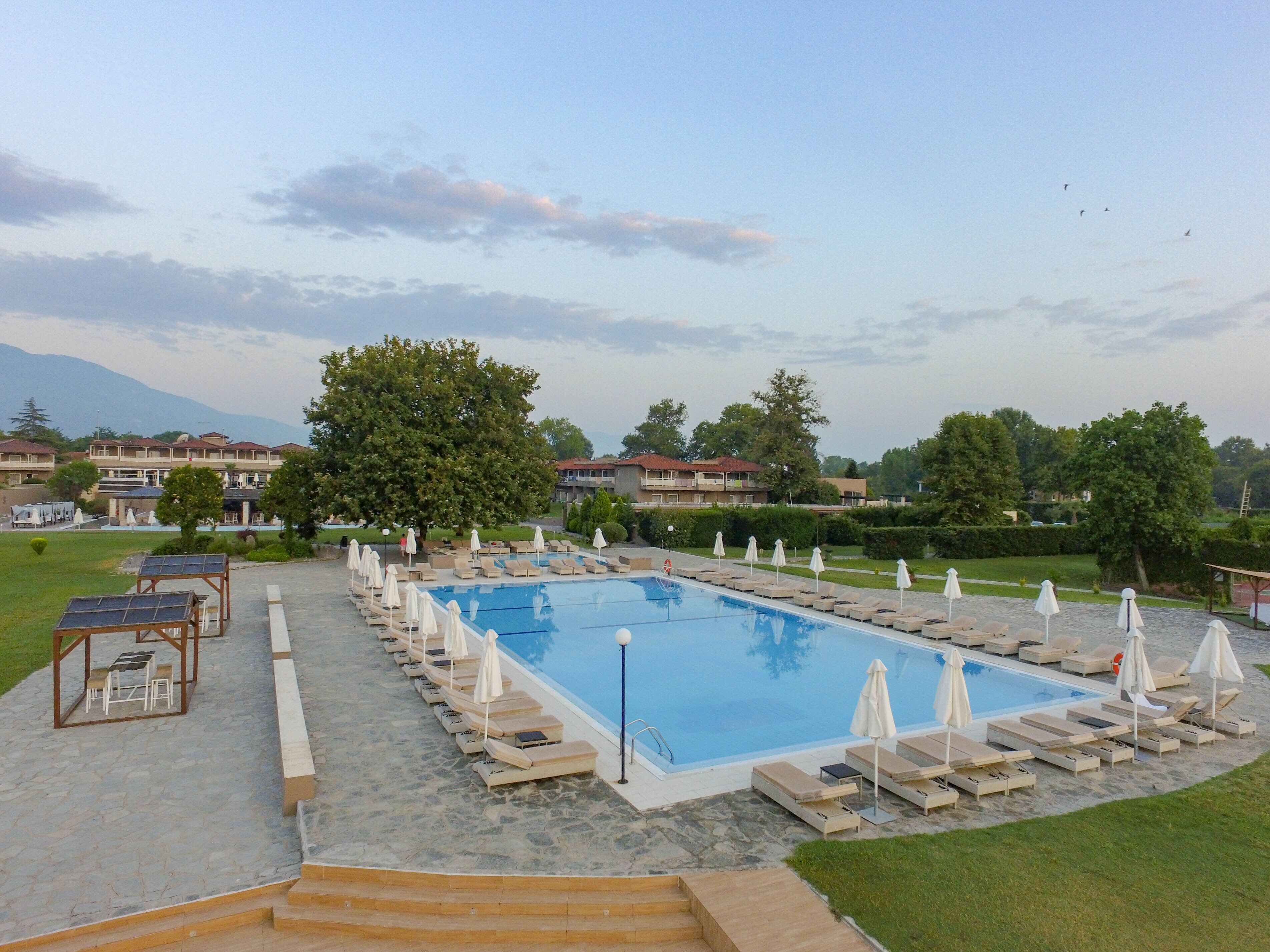 A swimming pool with sun loungers and umbrellas, surrounded by a stone deck and a view of buildings and trees.