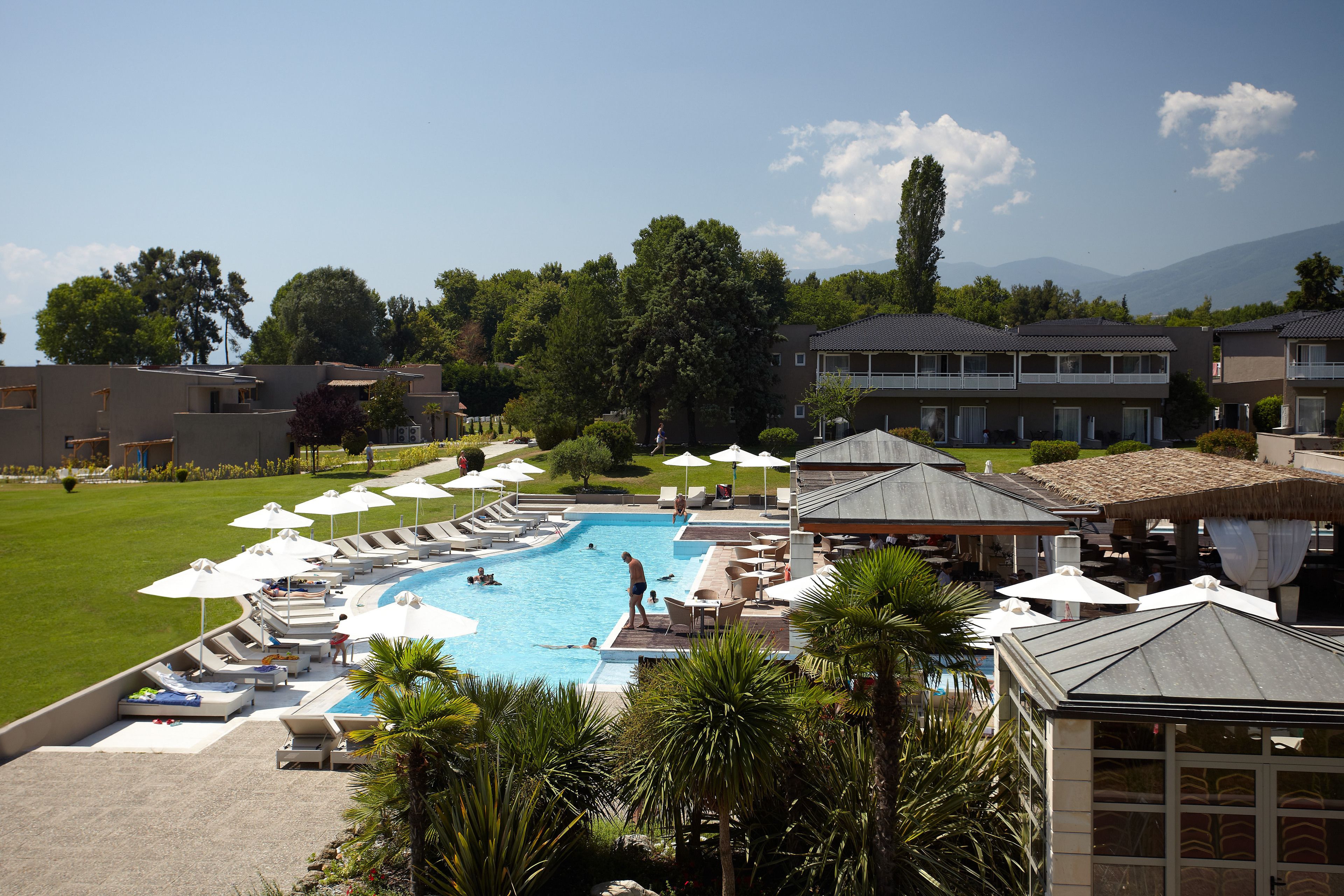 A resort with a large swimming pool surrounded by lounge chairs and umbrellas, with buildings and trees in the background.