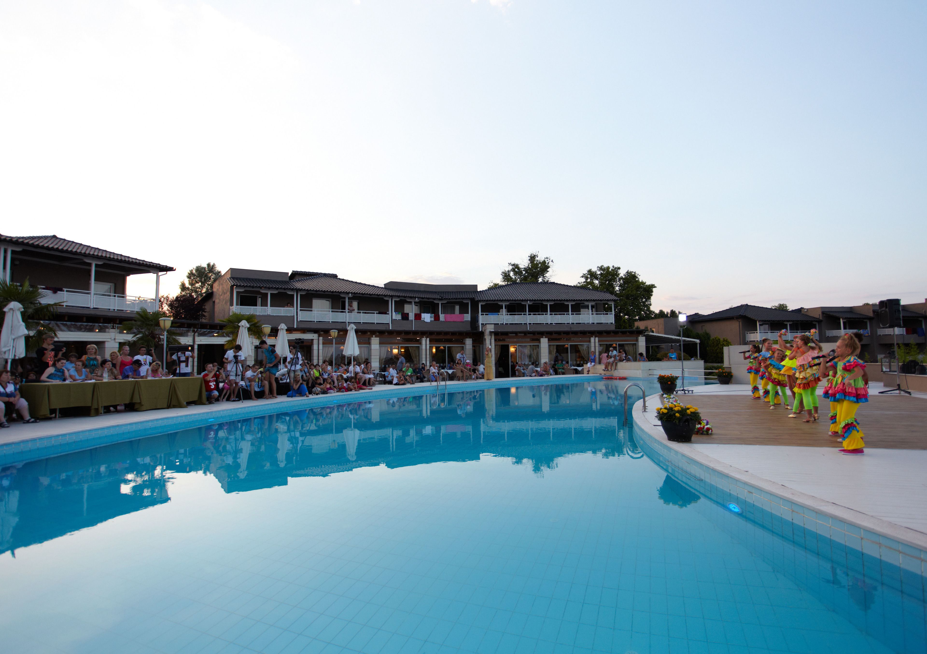 A swimming pool with people around it, a covered area with tables, and buildings in the background.