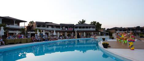 A swimming pool with people around it, a covered area with tables, and buildings in the background.