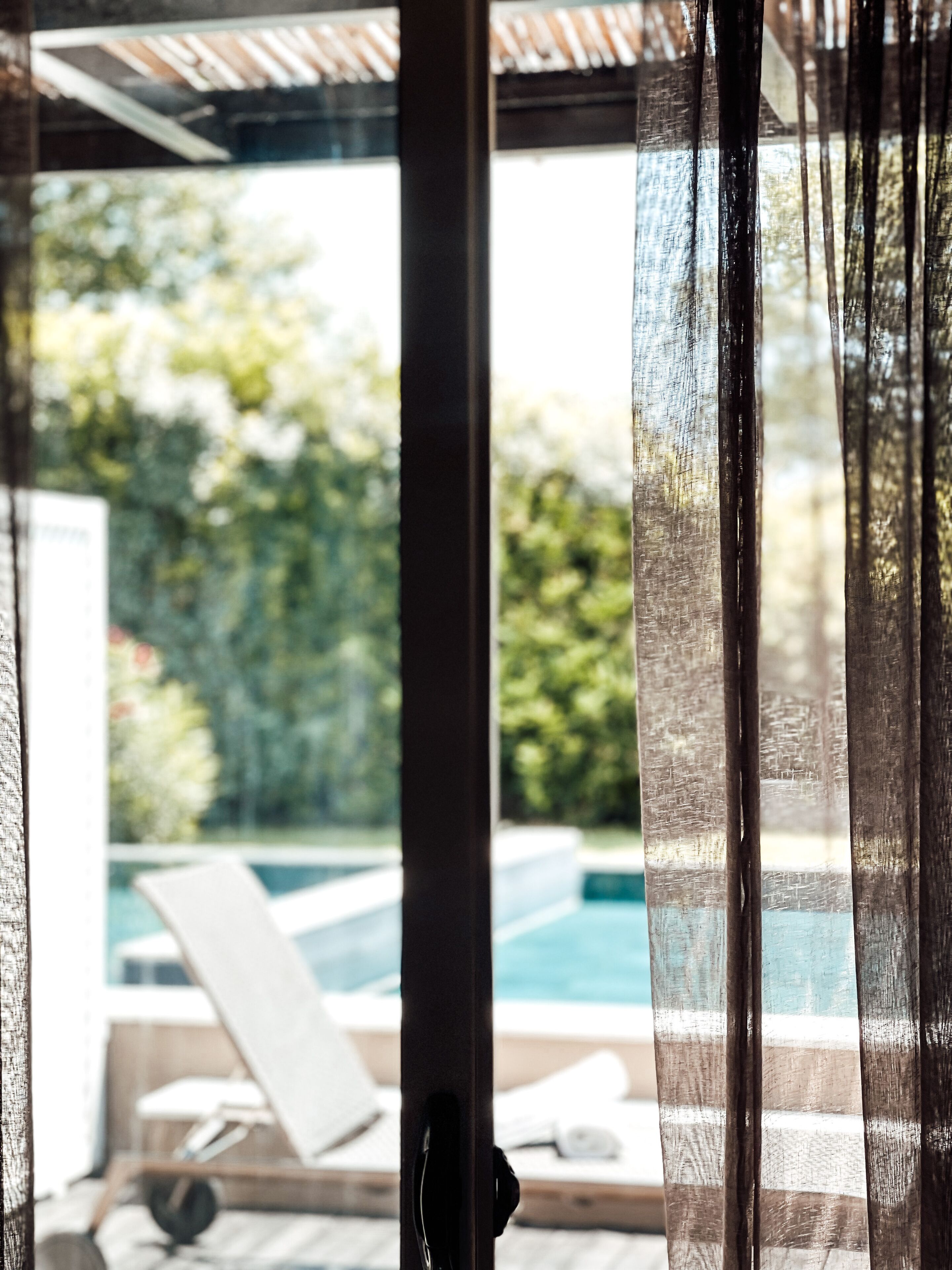 A view through a window with sheer curtains, revealing a pool area with lounge chairs and a wooden deck.