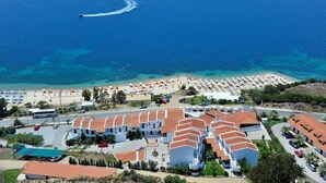 Vista aérea de un balneario costero con varios edificios, una playa con sombrillas y tumbonas, y un mar azul cristalino.