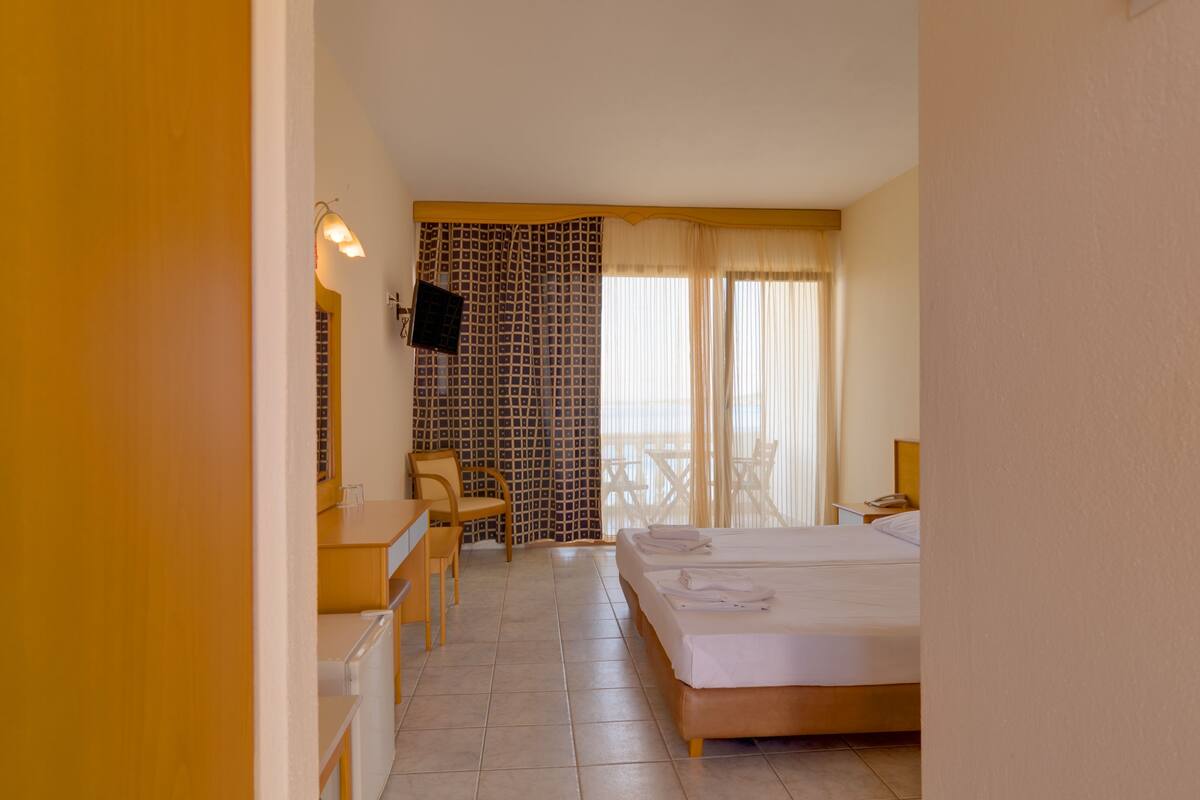 A hotel room with a bed, a desk, a chair, a TV, and a balcony with curtains.