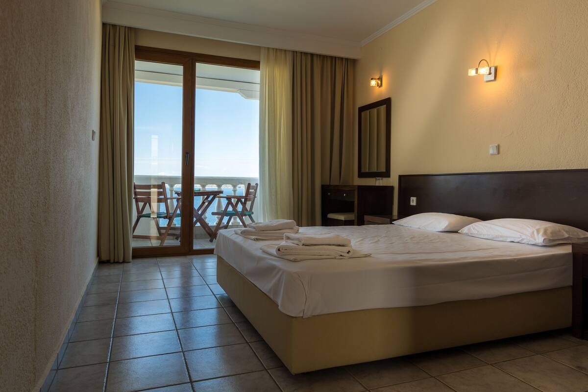 A hotel room with a bed, a chair, a table, a balcony with a view, and a wall-mounted light fixture.