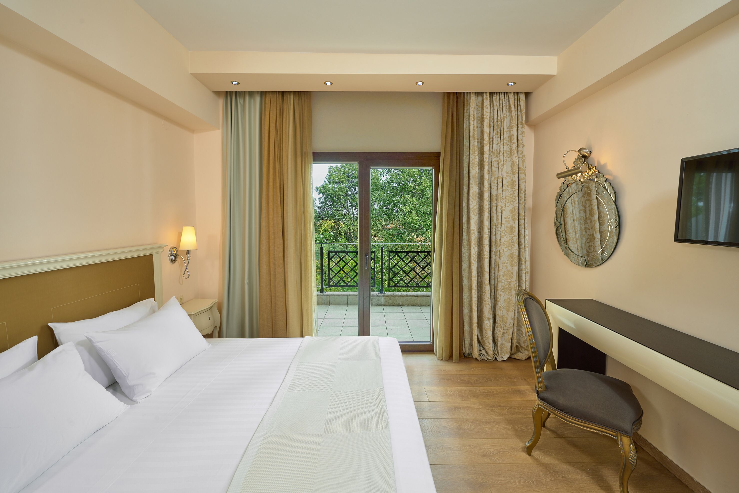 A hotel room with a bed, a desk, a chair, a TV, and a balcony with a view of trees.