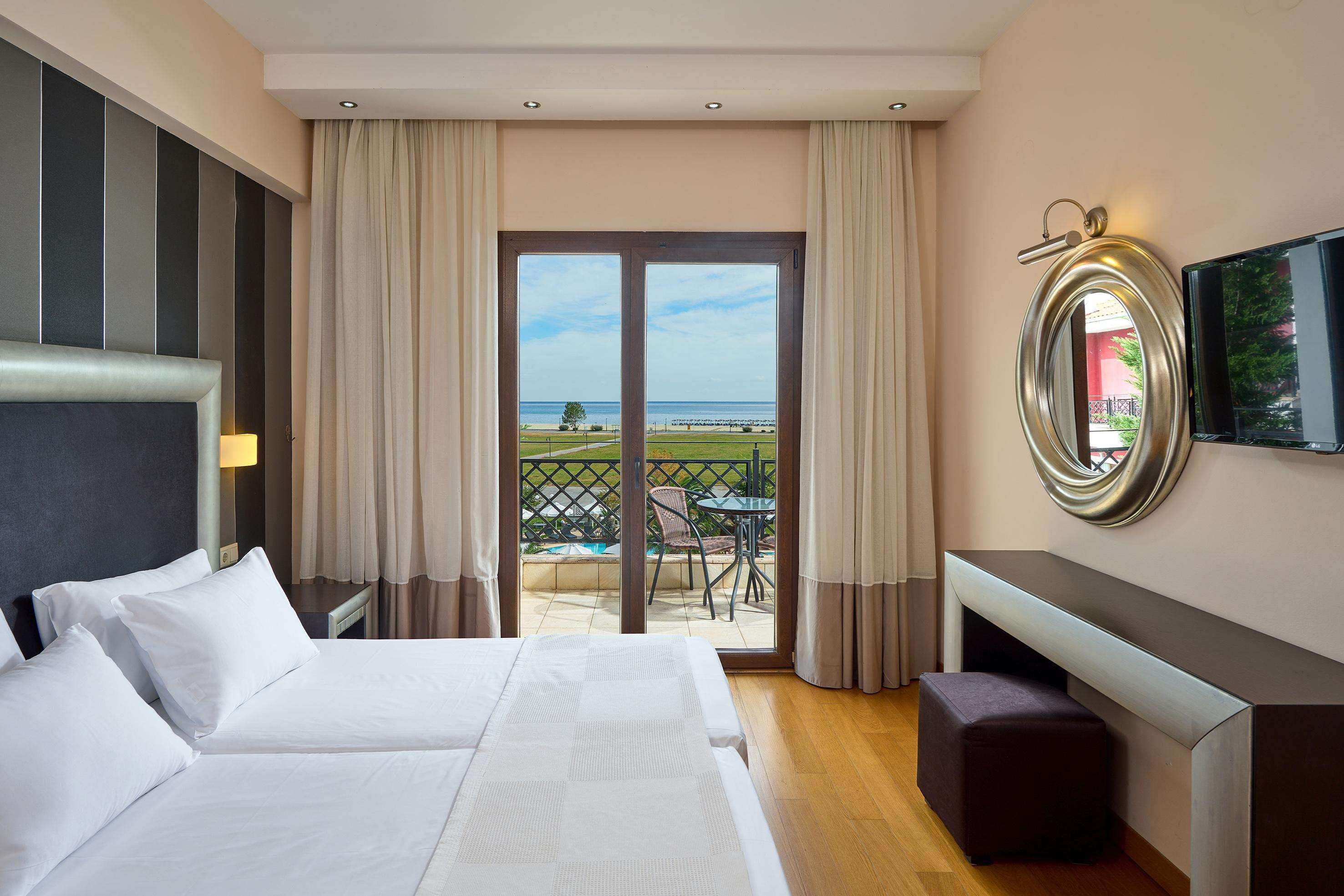 A hotel room with a bed, a desk, a TV, and a balcony with a view of the sea.