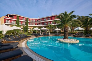 A hotel with a swimming pool surrounded by sun loungers and umbrellas, palm trees, and a red building facade. - Mediterranean Princess - Adults Only (Katerini)