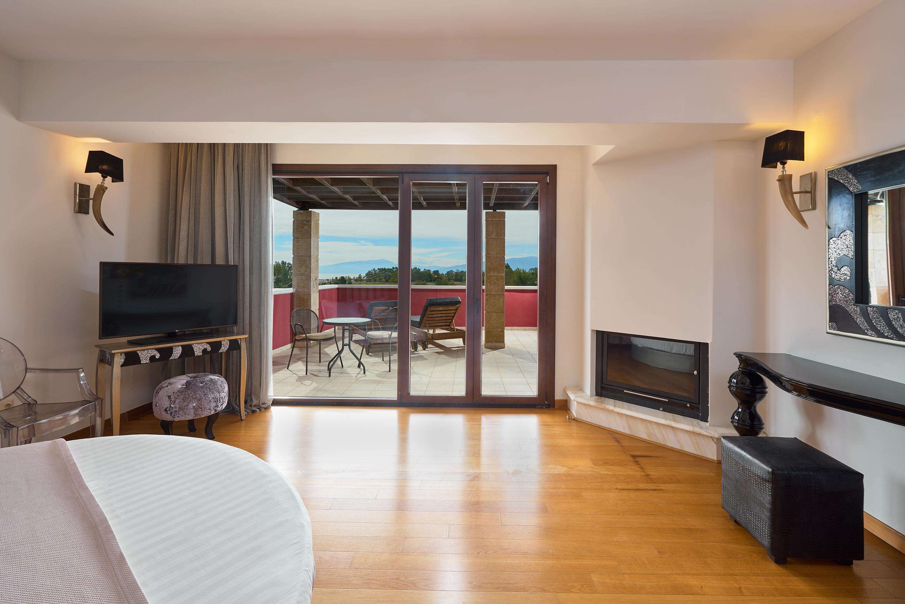 A modern hotel room with a TV, a fireplace, a balcony with a table and chairs, and a large window offering a scenic view.