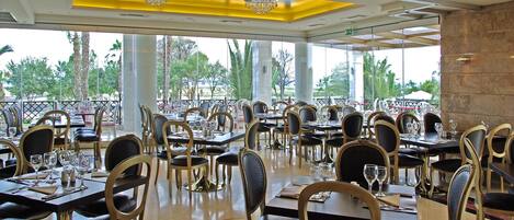 A spacious restaurant with multiple tables and chairs, large windows, and a chandelier.