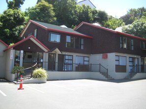 A two-story building with a red roof and brick exterior. There is a reception area with a sign, a staircase, and a small garden with a plant and a traffic cone. - 755 Regal Court (Dunedin)