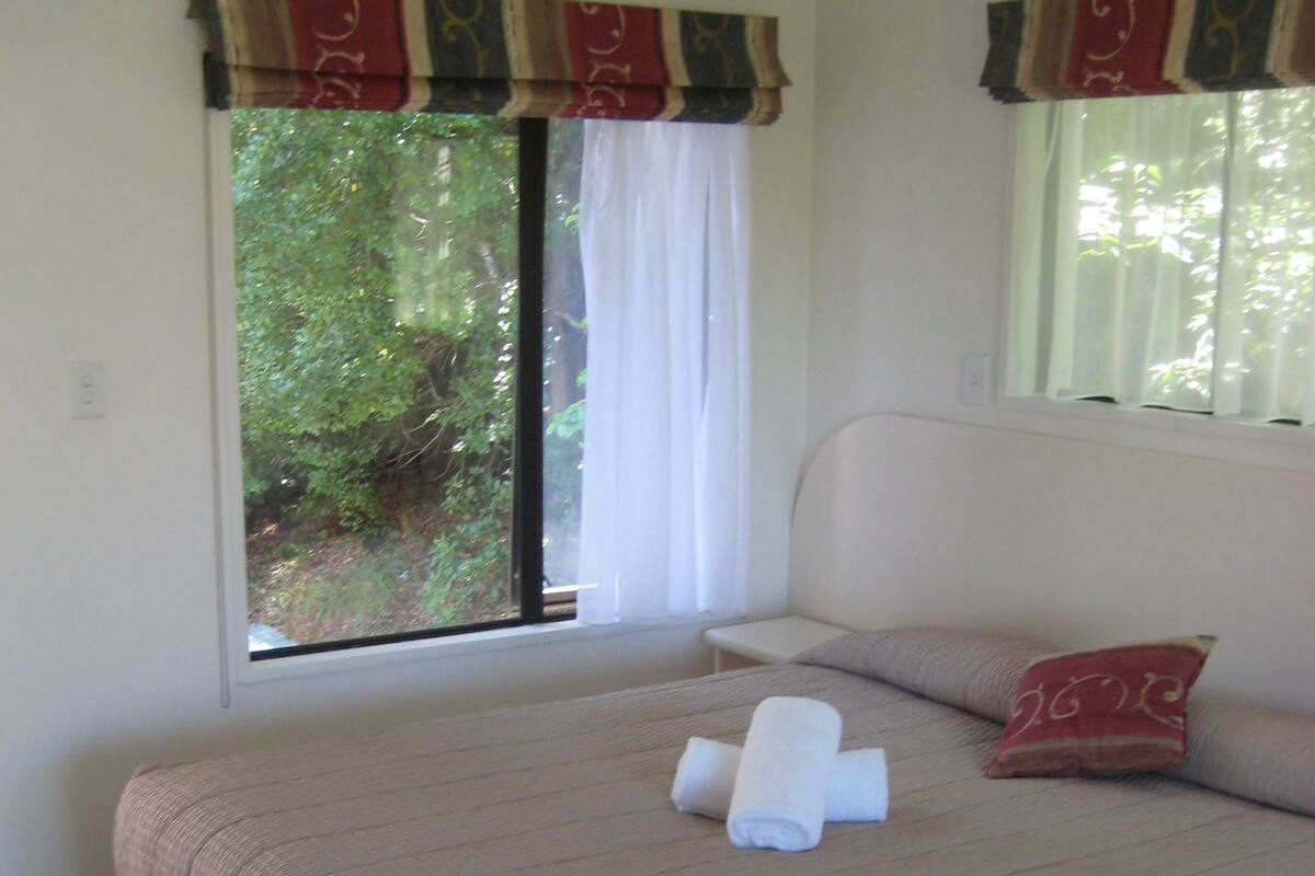A neatly made bed with a beige comforter, two rolled towels, and a red pillow.