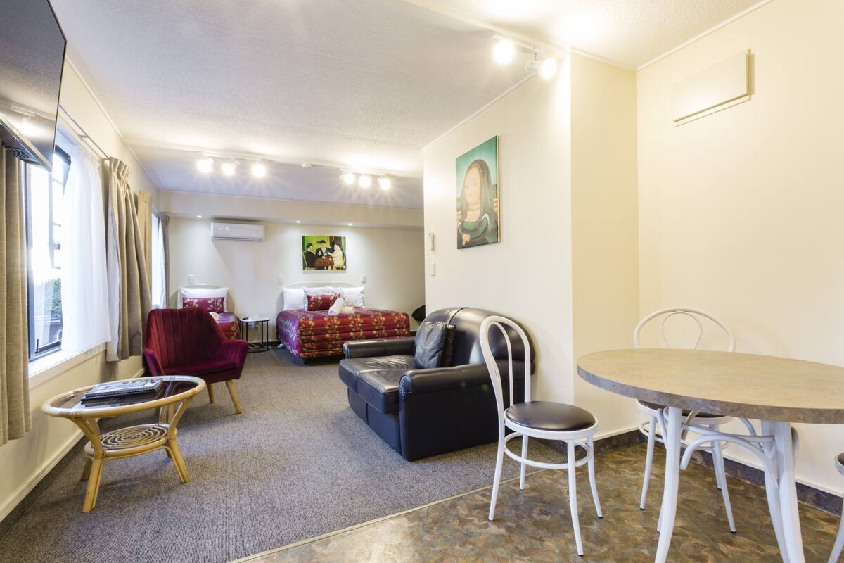 A hotel room with a bed, a sofa, a dining table, and a chair. There is a window with curtains, a painting on the wall, and an air conditioning unit.