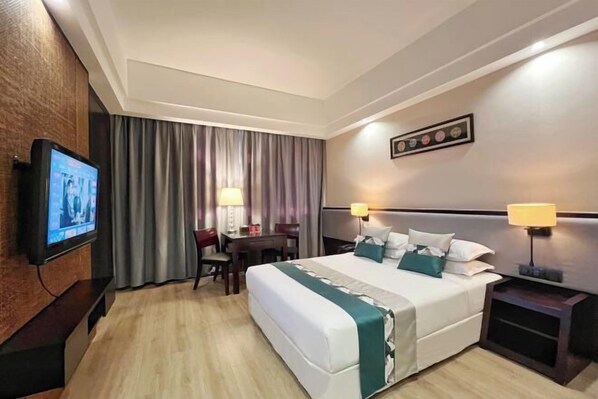 A hotel room with a large bed, two bedside tables with lamps, a desk with a chair, a television on the wall, and a framed picture on the wall.