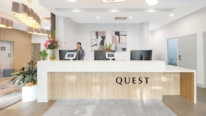 En modern receptionsdisk med ordet ”QUEST” tydligt synligt. Det finns två datorskärmar, en krukväxt och en vas med blommor. Receptionen har en stilren och minimalistisk design med en trädetalj på disken och en mönstrad matta på golvet.
