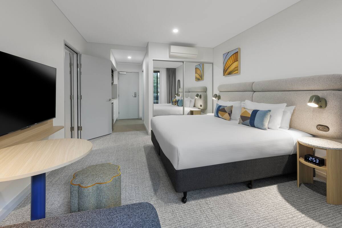 A modern hotel room with a large bed, a flat-screen TV, a bedside table, and a small lamp.