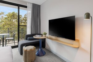 Una habitación de hotel moderna con un televisor grande montado en la pared, un espacio de descanso cómodo con un sofá y una mesa redonda pequeña, y un balcón con vistas a árboles.
