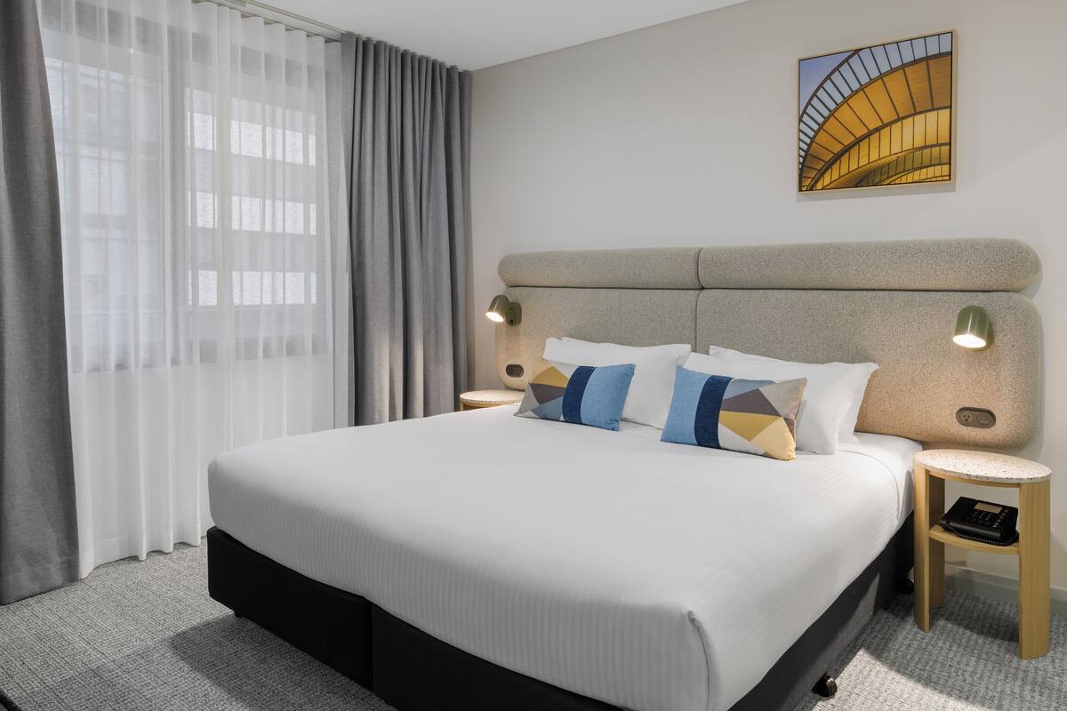 A modern hotel room with a large bed, two bedside tables, a small round table, and a wall-mounted artwork.