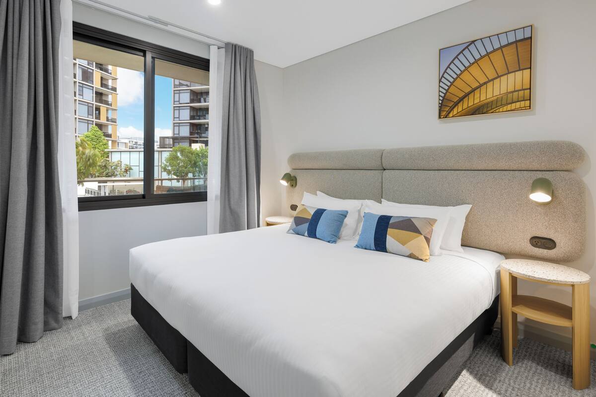 A modern hotel room with a large bed, a view of the city, and a small side table.