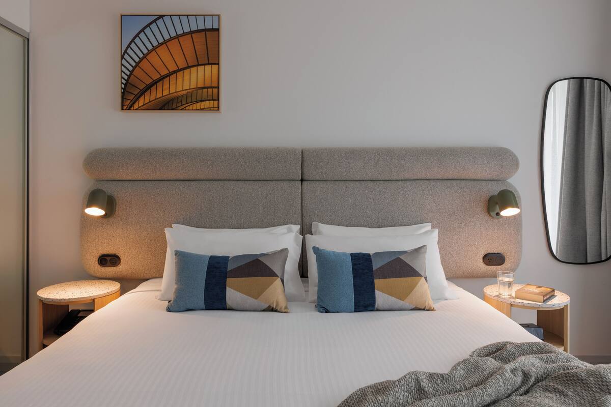 A modern hotel room with a large bed, two bedside tables, a wall-mounted artwork, and a mirror.
