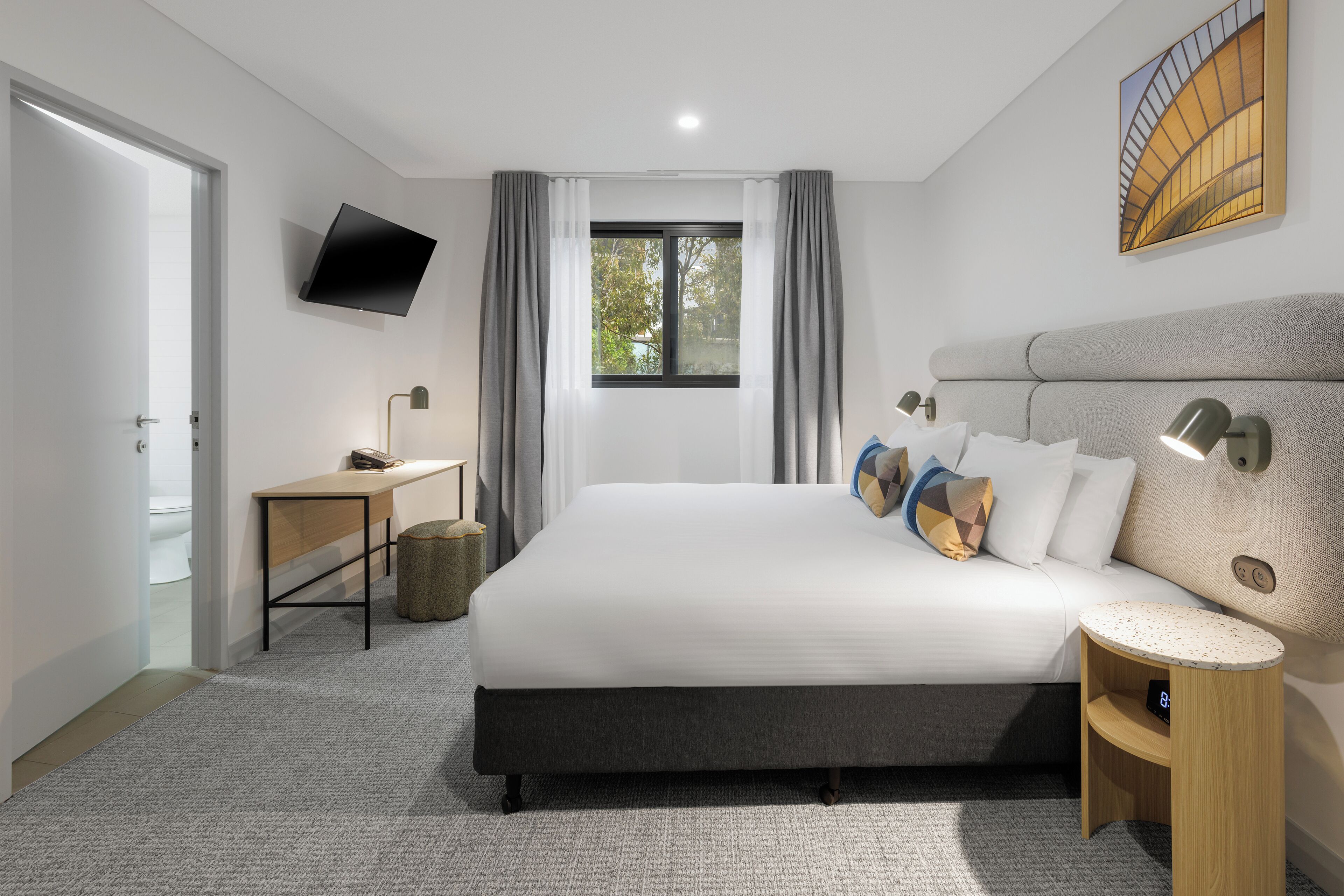 A modern hotel room with a large bed, a desk, a flat-screen TV, and a view of the outdoors.