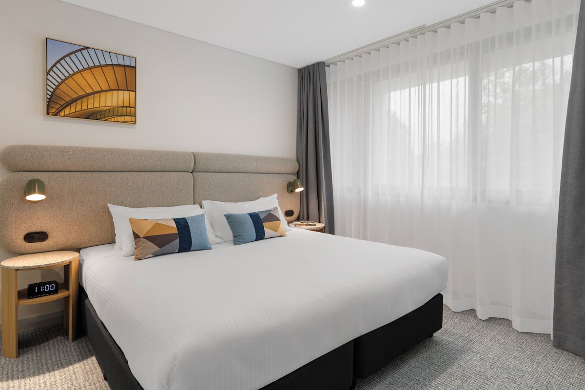 A hotel room with a large bed, two bedside tables, a wall-mounted clock, and a window with sheer curtains.