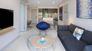 A modern living room with a flat-screen TV, a sofa, a round coffee table with a book, a blue chair, and a kitchen in the background. - Quest At Sydney Olympic Park (Sydney Olympic Park)