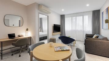 A hotel room with a desk, chair, round table, sofa, TV, and a large window with curtains.