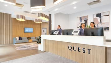 A modern reception area with a wooden reception desk labeled 'QUEST,' a flat-screen TV, and a seating area with a sofa and a coffee table.
