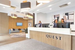 A modern reception area with a wooden reception desk labeled 'QUEST,' a flat-screen TV, and a seating area with a sofa and a coffee table. - Quest At Sydney Olympic Park (Sydney Olympic Park)