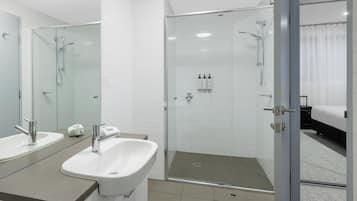 A modern bathroom with two sinks, a glass shower enclosure, and a visible bedroom with a bed and curtains.