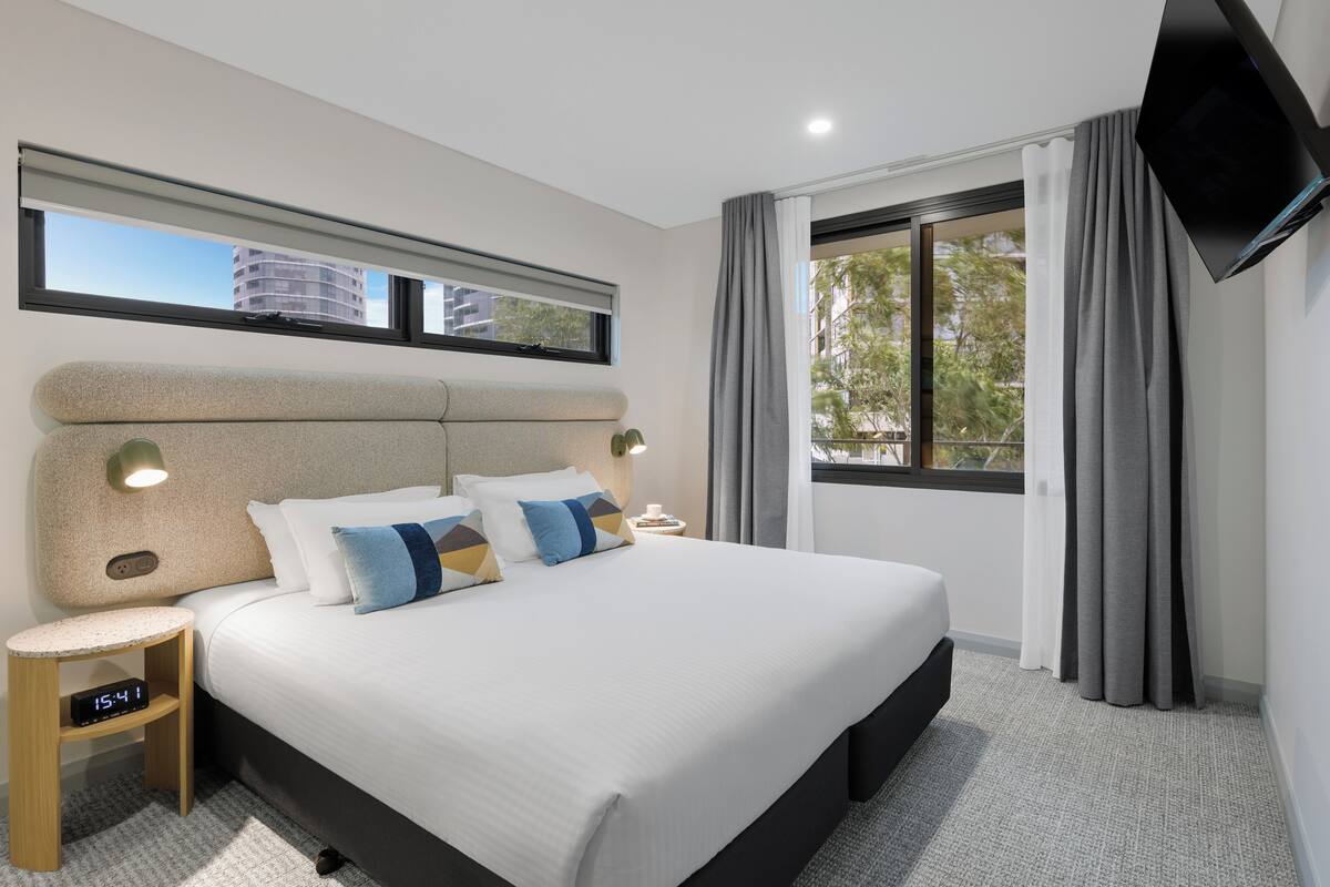 A modern hotel room with a large bed, a bedside table, a TV mounted on the wall, and a window with curtains.