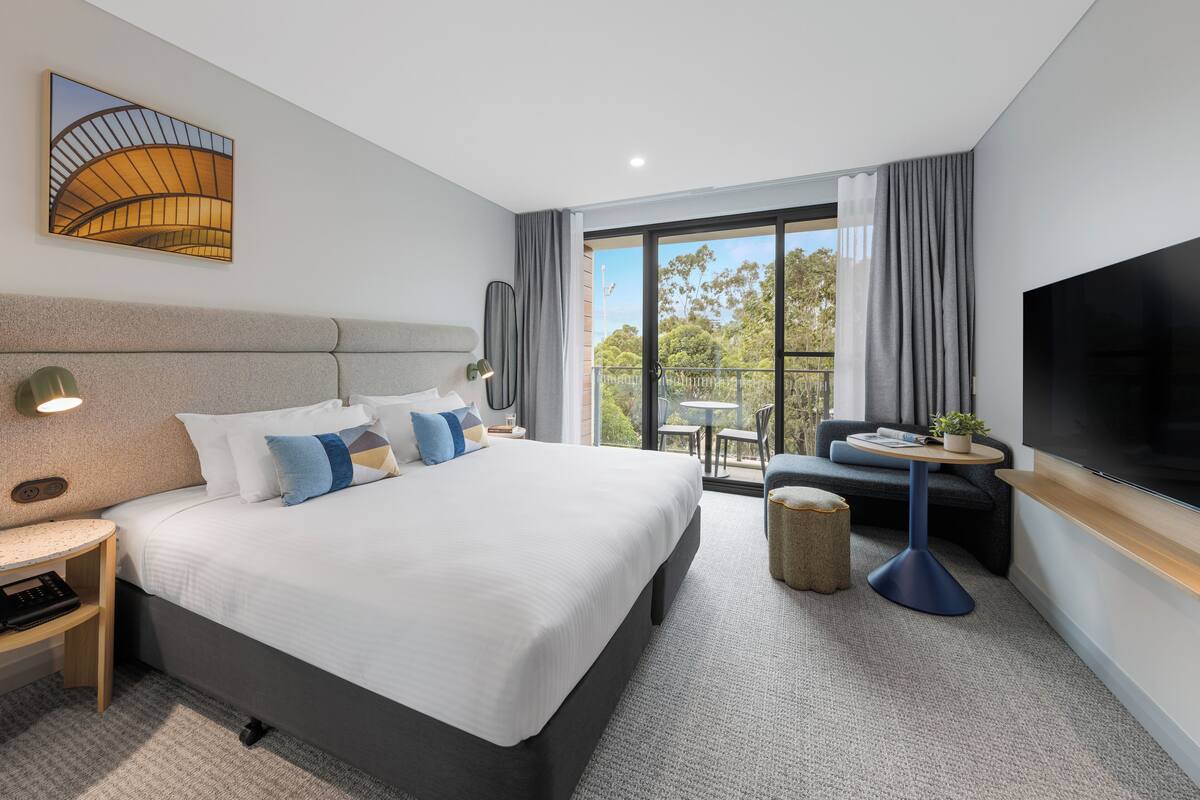 A modern hotel room with a large bed, a flat-screen TV, a balcony with a view, and a small table with a plant.